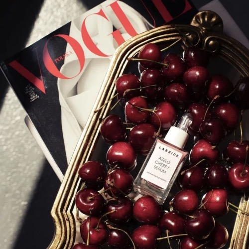 Szklana buteleczka z pipetą serum LABSIDE Azelo Cherry Serum leżąca na ciemnoczerwonych czereśniach w ozdobnej, złotej tacy. W tle widoczny jest fragment magazynu Vogue.